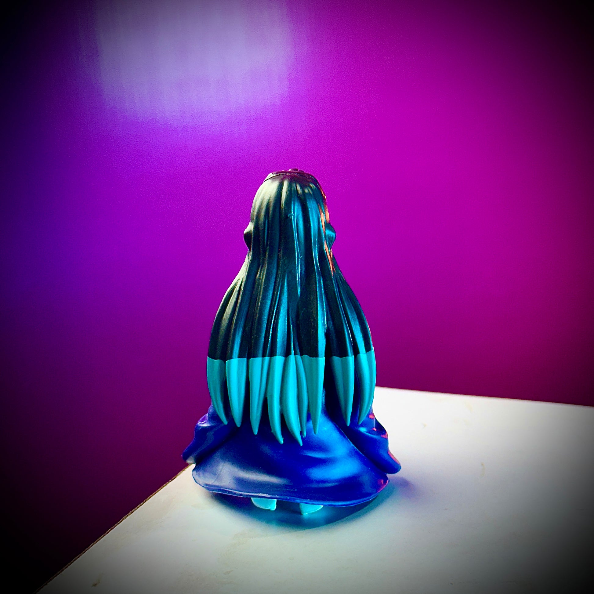Blue and black figurine with long hair on a white surface against a purple background